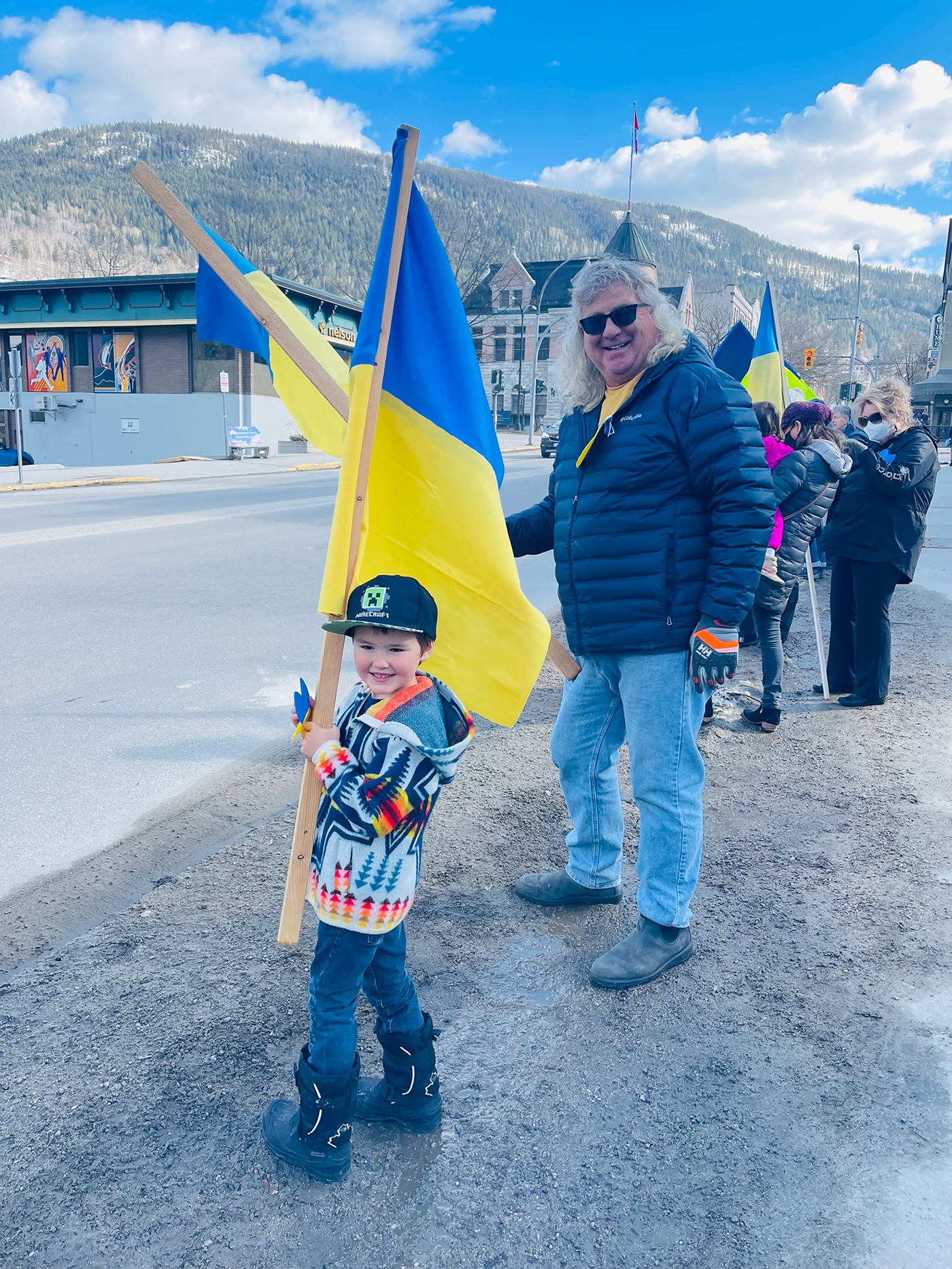 A child in a colorful sweater holds a Ukrainian flag, while an adult beside him smiles and also holds a flag. They are outdoors, with a group of people holding flags in the background and mountains visible.