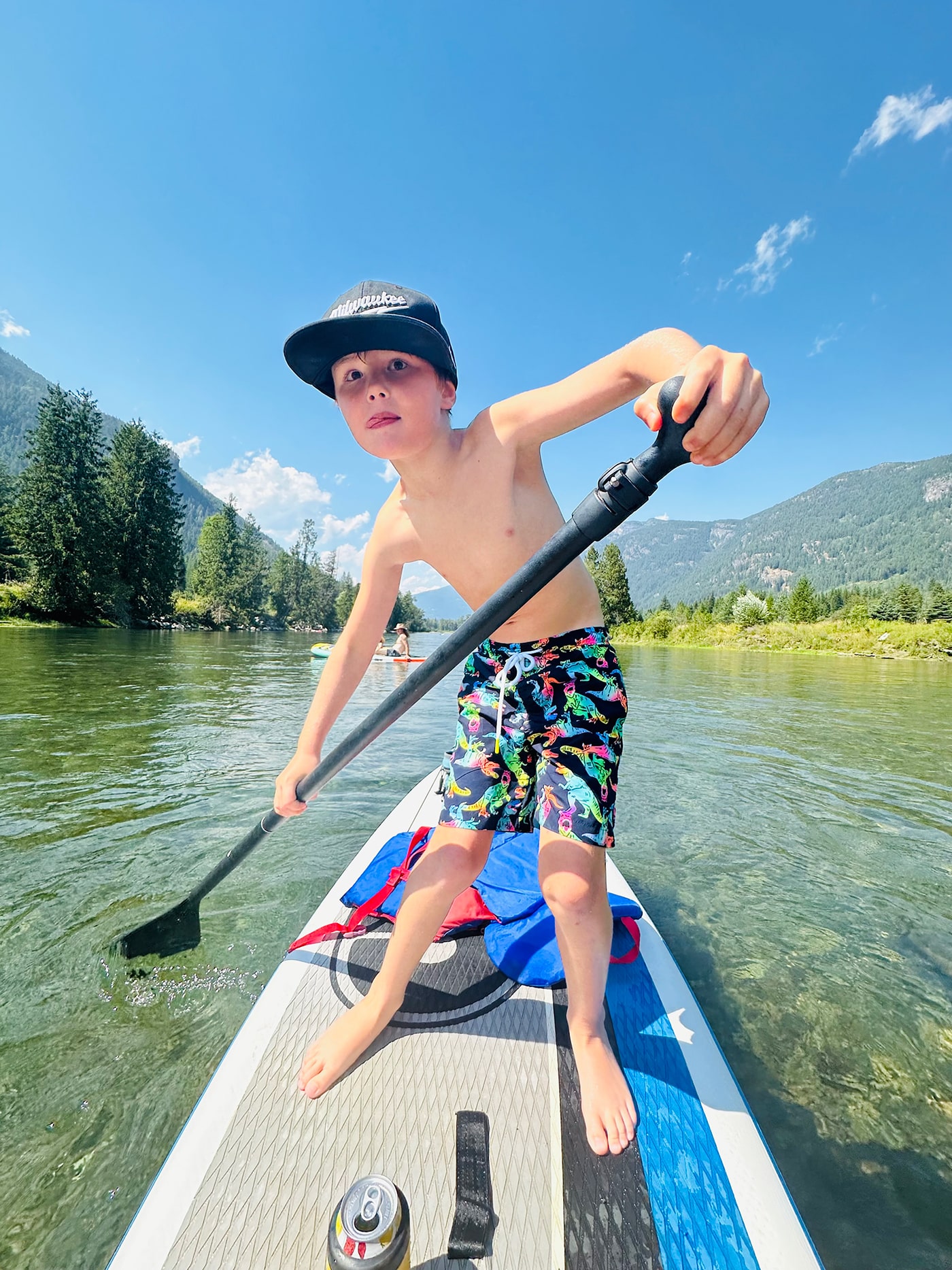 A young boy stands on a paddleboard in a river, wearing colorful swim shorts and a black cap, paddling with one hand while looking surprised. The background features trees and mountains under a clear blue sky.