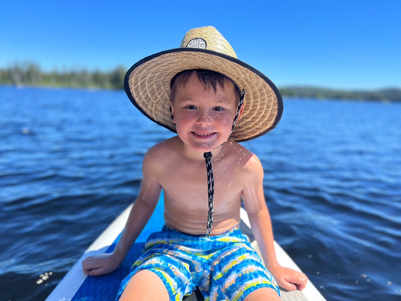 A young boy wearing a large straw hat is sitting on a paddleboard in a body of water, smiling at the camera, with sunlight reflecting on the water.