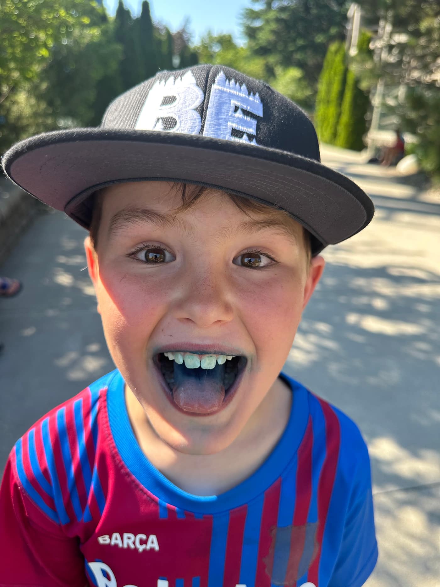A young boy is smiling widely, sticking out his tongue which is blue, and wearing a black cap with the word "BE." He is in a casual outfit and the background includes trees and a pathway.