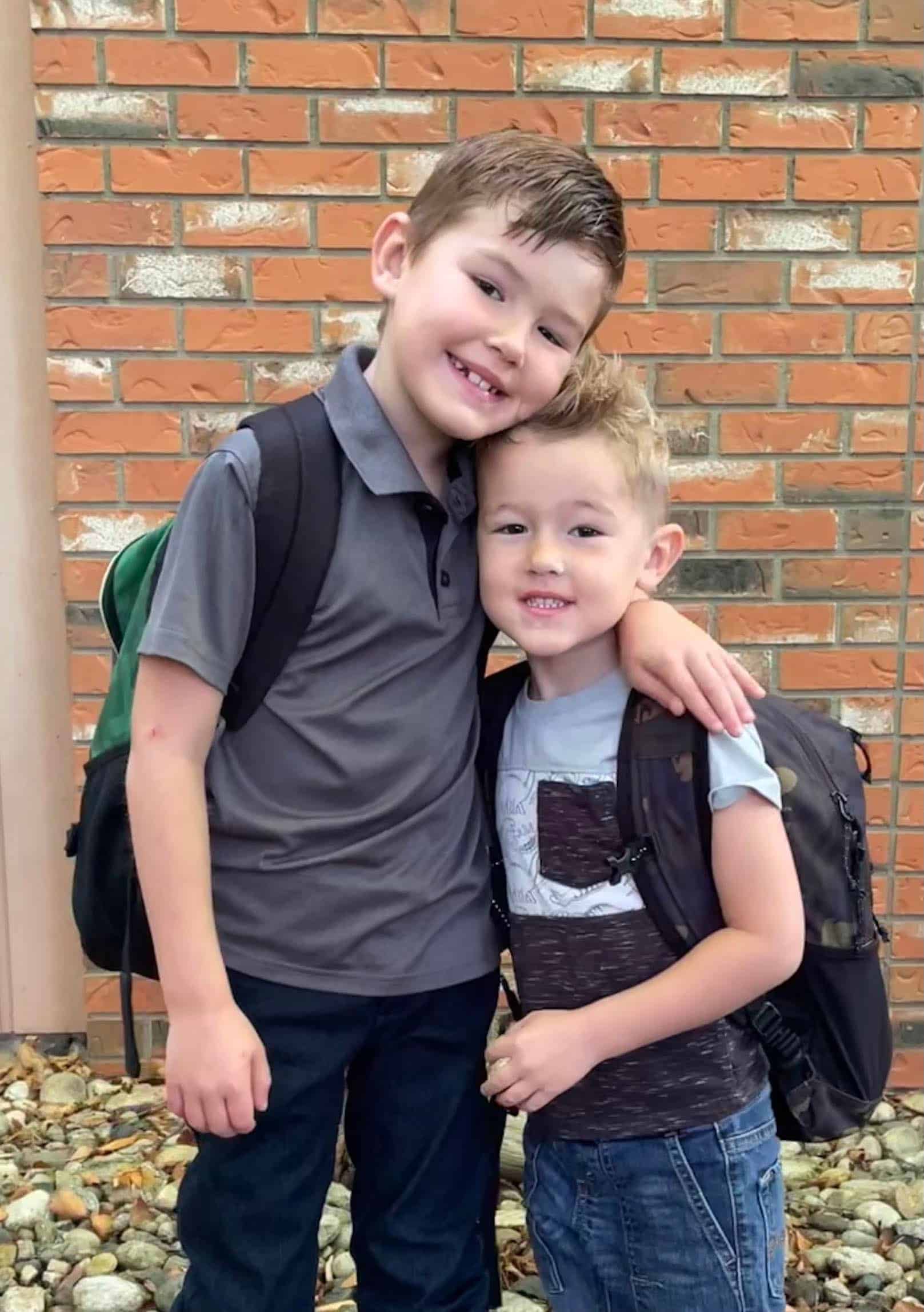 Two young boys standing close together, smiling, in front of a brick wall. They are wearing backpacks and casual clothes.