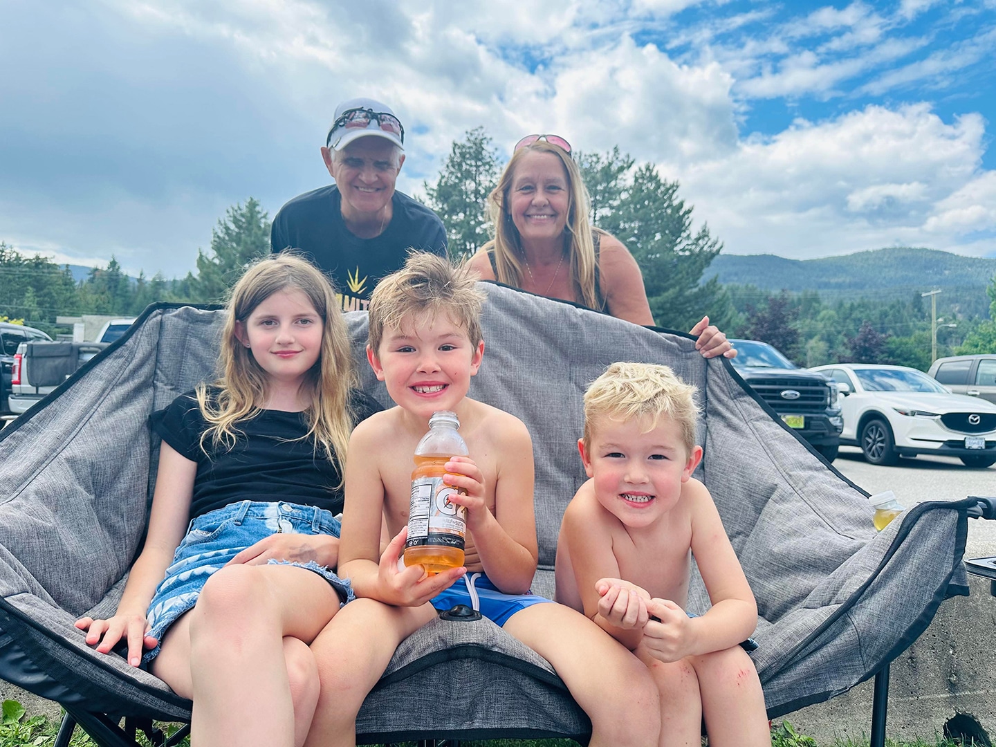 A family of five is enjoying a sunny day outdoors. Two girls and three boys sit on a large chair, with one boy holding a drink. There are trees and cars in the background.