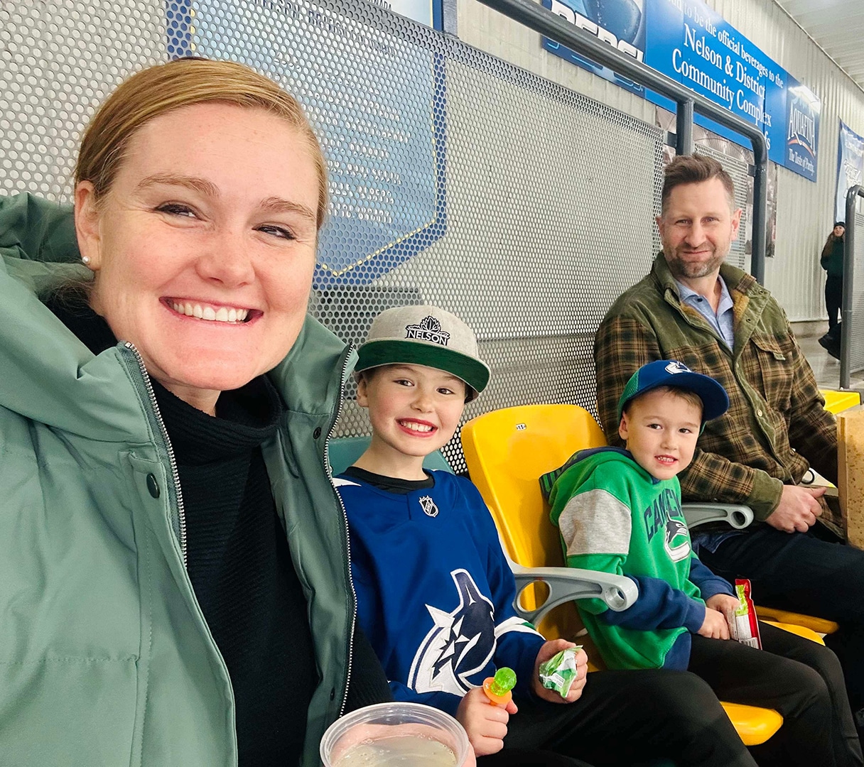 A smiling family sits together in a rink, wearing team jerseys and caps, enjoying snacks during a hockey game.