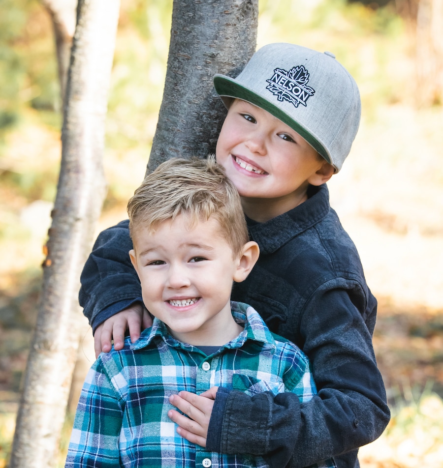 Two young boys posing together outdoors, one wearing a gray cap and the other in a plaid shirt, both smiling at the camera.
