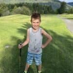 A smiling boy in casual clothing stands on a golf course, holding a golf club, with a green landscape and mountains in the background.