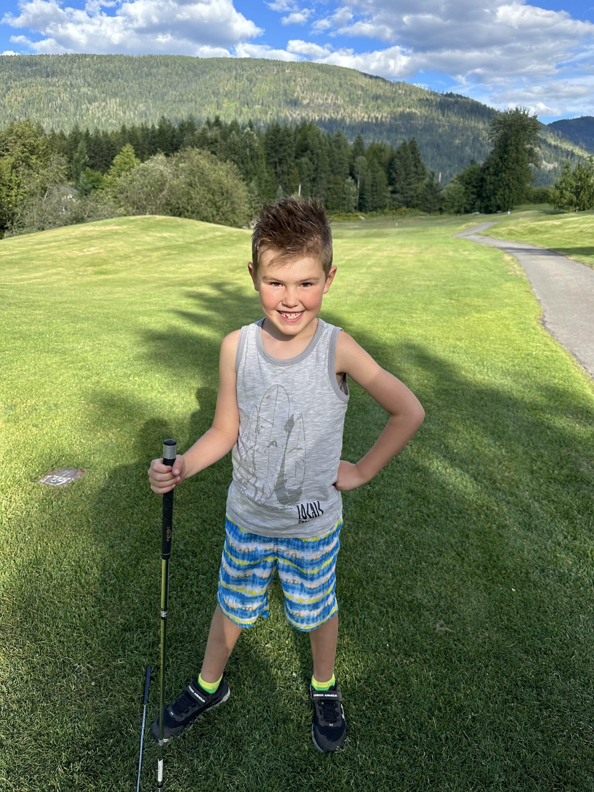 A smiling boy in casual clothing stands on a golf course, holding a golf club, with a green landscape and mountains in the background.