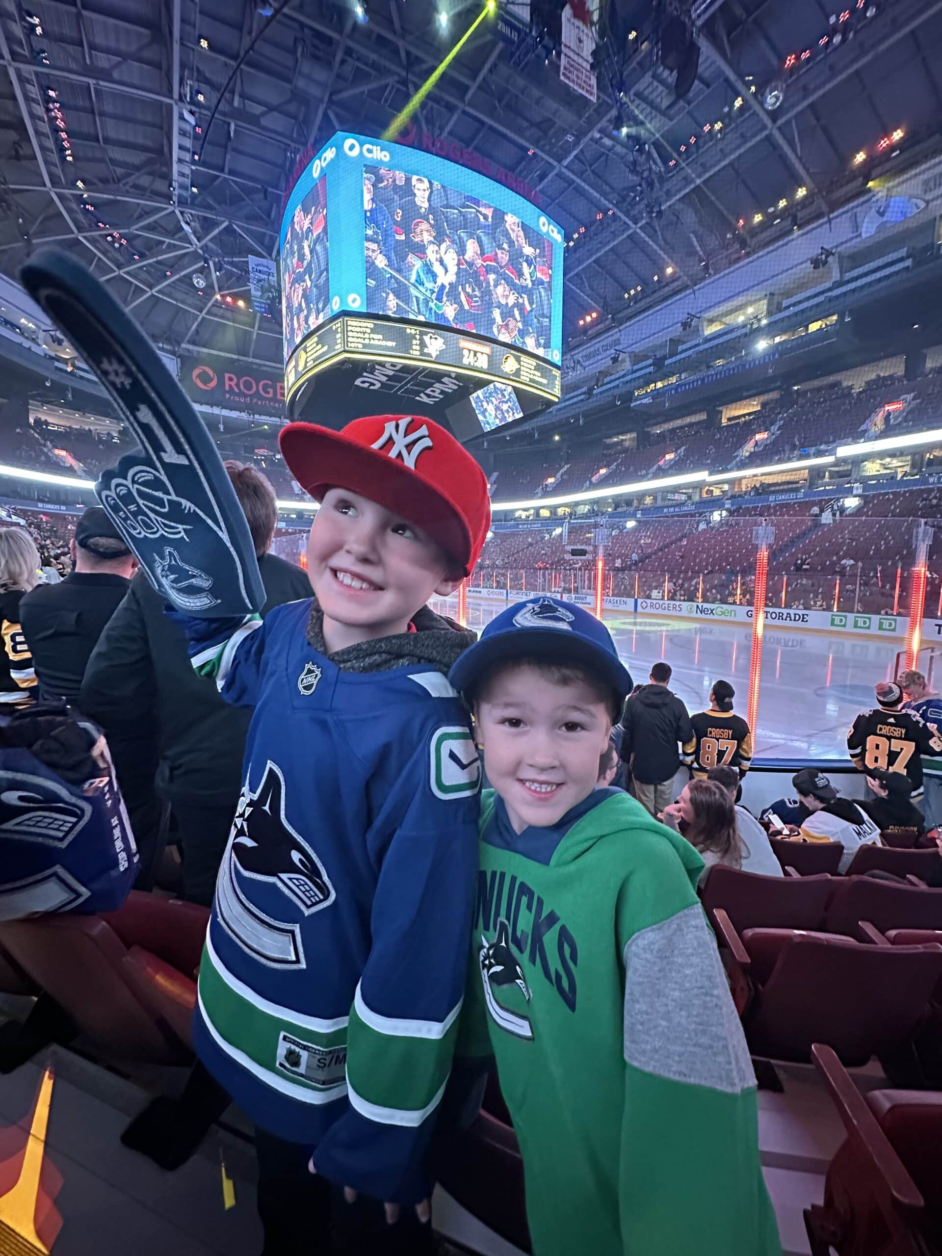 Two young boys at a hockey game, smiling and posing with team gear. One is wearing a Vancouver Canucks jersey and the other a green hoodie with "Canucks" printed on it. A big screen displaying the game is visible in the background.