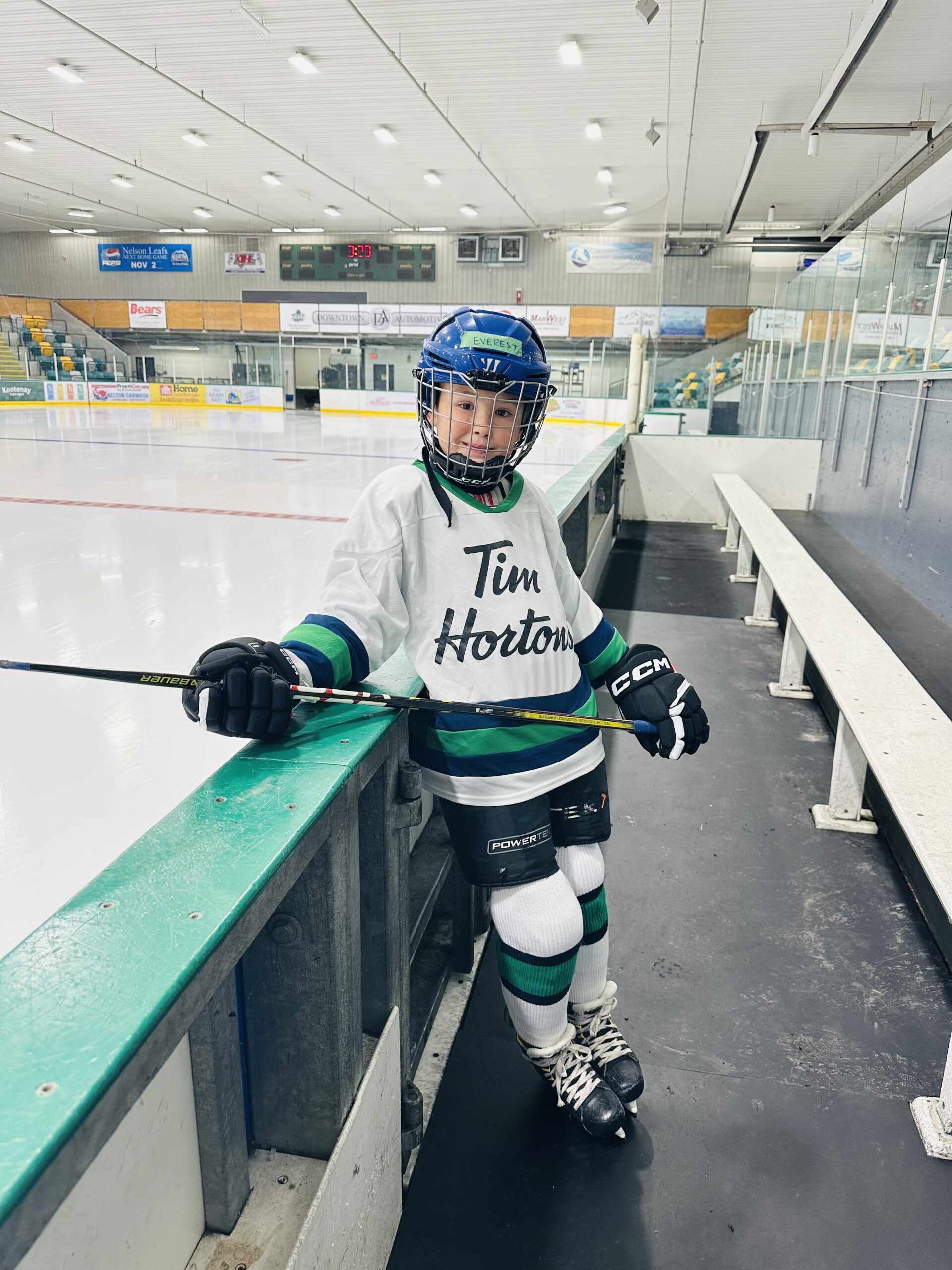 A young hockey player in a white "Tim Hortons" jersey leans against the rink barrier, wearing a helmet, gloves, and skates, with a hockey stick in hand, in an ice rink setting.