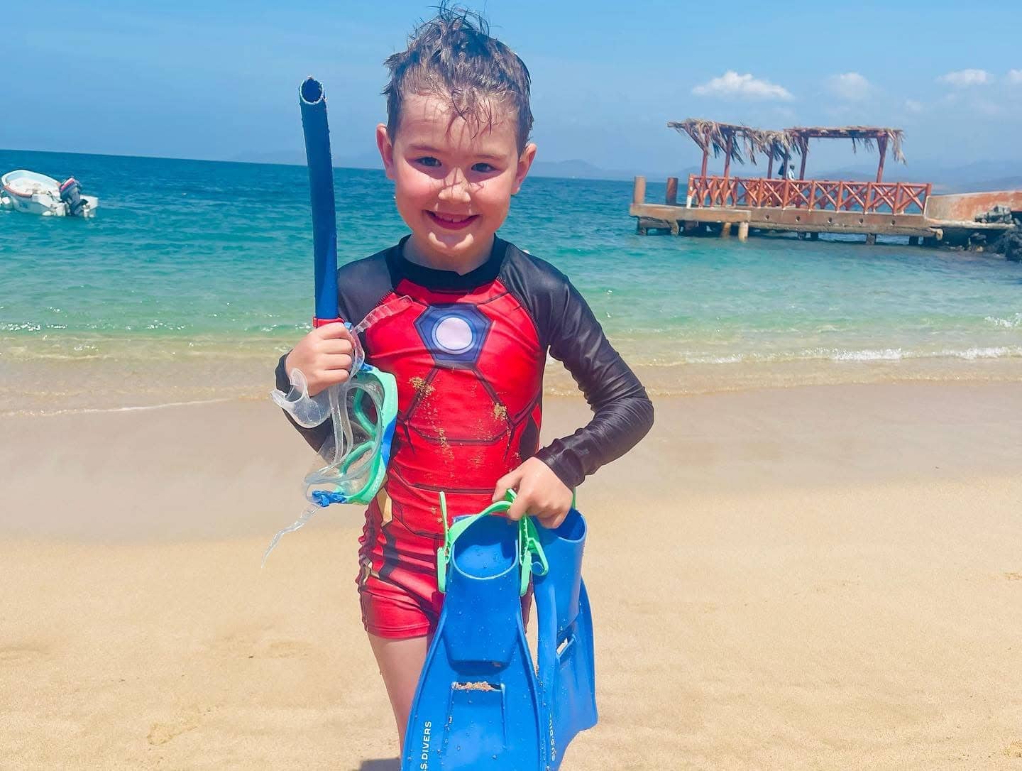A child wearing a red swimsuit with a black sleeve is standing on a beach, holding a snorkel and a pair of blue fins. The ocean and a wooden pier are visible in the background.