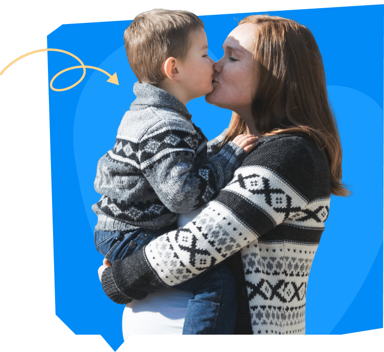A woman kisses a young boy while holding him. They are both wearing sweaters with patterns, and the background is bright blue.