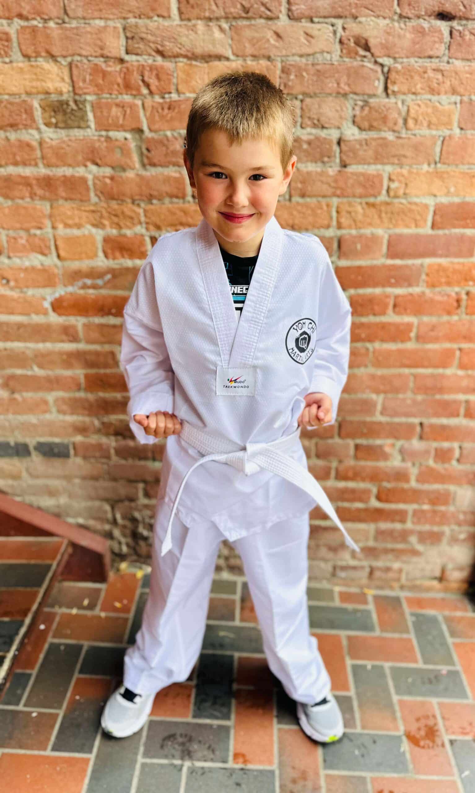 A smiling boy wearing a white martial arts uniform stands against a brick wall.